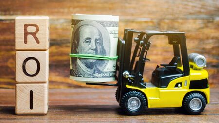 Wooden blocks with the word ROI and money with forklift. Ratio between the net profit and cost of investment resulting from an investment of resources. High level of business profitability.の写真素材