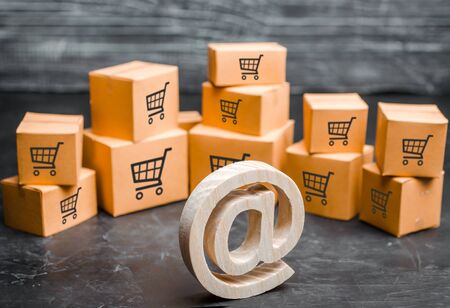 Wooden email symbol stands near the cardboard boxes. Concept of online trading and e-commerce. Distribution and retail. Shopping. Internet advertising. Marketing, targeting. Production of goodsの写真素材