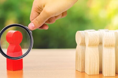 A magnifying glass looks at a red figurine of a man stands aside from the crowd of people. Asociality, sociopathy. Development of leadership and social qualities. Rejected from society, lonely.の写真素材