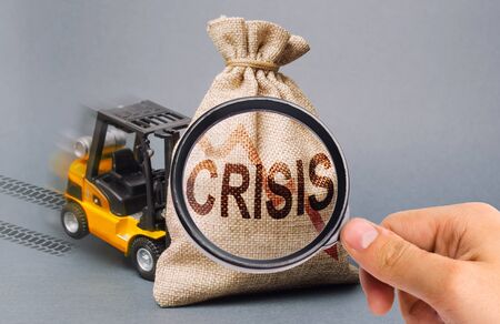 A magnifying glass looks at a yellow forklift truck can not lift the bag with the inscription crisis. Economic crisis, stagnation and recession of the economy. decline in production, purchasing power.の写真素材