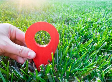 A hand is holding a red location marker in the green grass. The concept of tourism and travel. Navigation and exploration. Destination. Holiday, vacation. Buying building land. Selective focusの写真素材