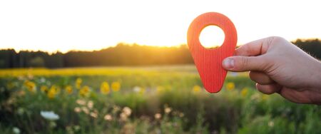 A hand is holding a red location marker in the sunset background. The concept of tourism and travel....