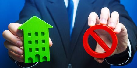 Man holds a green house and red prohibition symbol NO. Prohibition of operation. Denial of insurance payment. Bank refuses to provide a mortgage loan. Housing problems. Confiscation pledged property.の写真素材
