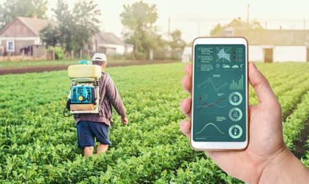 A hand with a phone on background of a farmer with agriculture smoke fog spraying machine. Crop...
