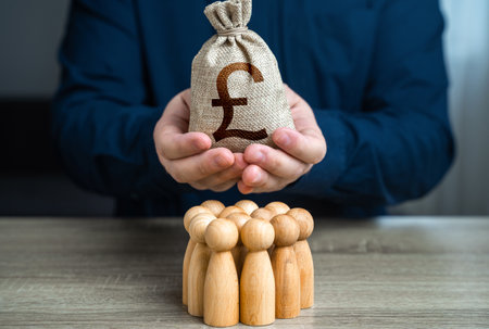 A man is holding a British pound sterling money bag over a group of people figurines. Political lobbying. Payment for labor and services. Generation of funds. Organization of the community budget.の写真素材