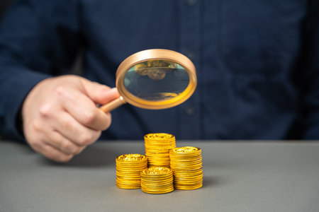 The concept of analyzing income and profit in a company. Business efficiency and investment. Fund, savings. A businessman holds a magnifying glass over a stack of coins.の写真素材
