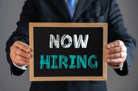 A businessman holds a sign that says Now Hiring. Job opportunities, recruitment, workforce expansion, employment growth, career openings, labor market demand, hiring trends, and business development.の写真素材