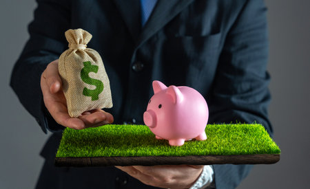 A businessman hands a dollar money bag to a piggy bank that is on the grass. Saving money, investment strategies, wealth accumulation. Smart budgeting, and long-term financial planning.の写真素材