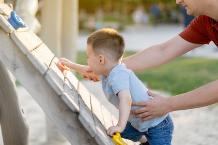 A cute happy caucasian toddler boy of three or four years old climbs a artificial climbing rock with holders and ledges for children on the playground with the dad. Spending time with family.の写真素材