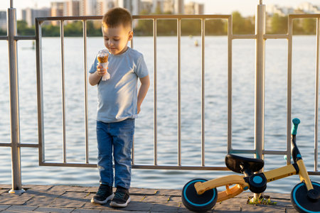 A Caucasian toddler boy of three and a half years old eats ice cream in a city park near a lake. Happy childhood concept. Sunny summer day. Cycling or bicycleの写真素材