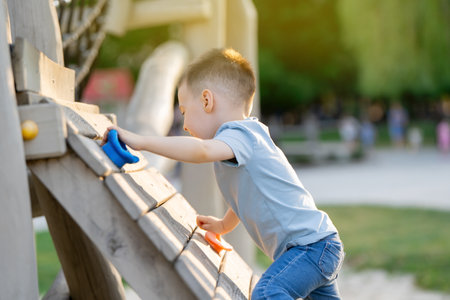 A cute happy caucasian toddler boy of three or four years old climbs an artificial climbing rock with holders and ledges for children on the playground. Spending time with family.の写真素材