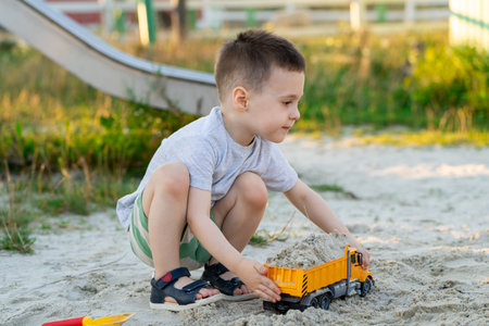 Little cute caucasian toddler boy of three and a half years old plays in the sandbox on a sunny summer day. Outdoor creative activities for kids. Toy dump truck and shovel.の写真素材