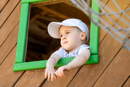 Pensive toddler boy of three or four years looks out of a window on a wooden playground. Toddlerhood and childhood concept. Active lifestyleの写真素材