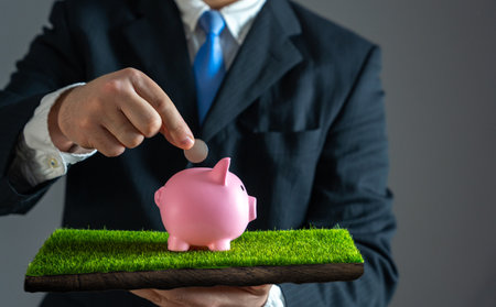 Businessman puts coin in piggy bank standing on grass. Concept of saving and investment and savings. Personal finance, budgeting. Financial growth. Wealth accumulation, smart money management.の写真素材