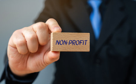 Block with the word non-profit. Organization that operates for a purpose other than generating profit for its owners or shareholders. Businessman holding wooden block in his handsの写真素材