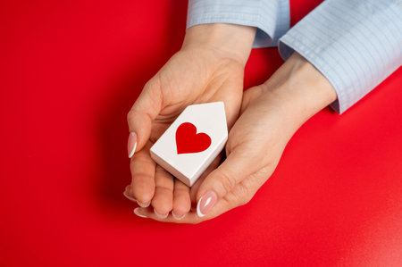 A house with a heart in the hands of a women. The concept of a love nest and the beginning of a life together. Relationship milestones, emotional security. Loving home, family bondsの写真素材
