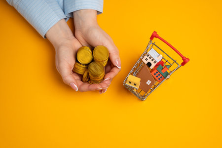 Concept of buying new housing and investing in real estate. Rent and mortgage for apartments. Affordable housing. Supermarket cart with houses and coins in a woman's handの写真素材