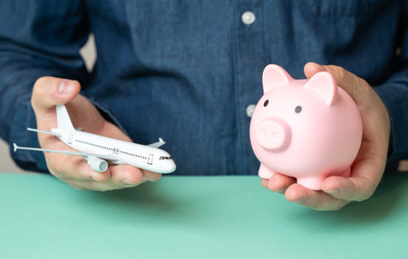 A man holds an airplane and a piggy bank. Travel savings. Budget flights. Cheap tickets. Cost optimization. Vacation planning. Tourism on a budget.の写真素材
