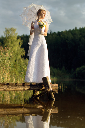 Beautiful bride with an umbrella on a bridge near the water in the forestの写真素材