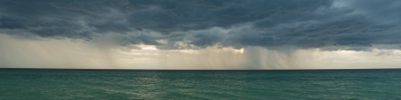 Panorama of dark stormy clouds over blurred sea water with sunset rays in cloud and rain threads 