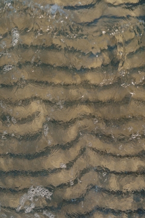 Texture of wave passing over sand ripples on sea bottom with clear transparent water bubbles and shadows from side lightの写真素材