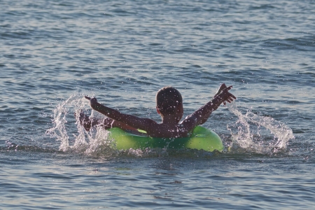 Boy lies in green inflatable circle in calm sea water and plays with splashes by his handsの写真素材