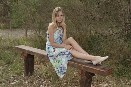 Beautiful young blond woman relaxing on wooden bench looking into camera with her legs lying on the benchの写真素材