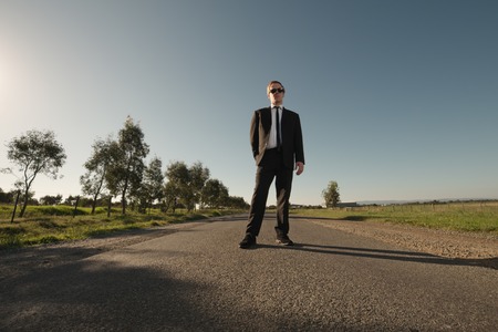 Businessman in sunglasses and black suit stands on empty asphalt road in wide angleの写真素材