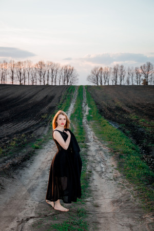 Girl in black dress is standing on the field  barefoot. woman outdoors. Sunset.の写真素材