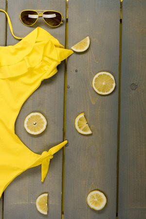 Woman beach accessories on wooden background. Yellow swimsuit one-piece, aviator sunglasses between parts of lemons on grey wooden board.の写真素材