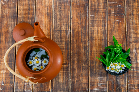 Open brown teapot with flower of chamomile near black plate with fresh mint and chamomile on wooden background. Frame. Copy space. Tea concept.の写真素材