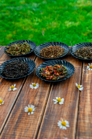Different kinds of tea on black plates on wooden table with flowers of chamomile in garden and on nature background. Assortment of dry tea. Tea concept. Tea leaves. Closeupの写真素材