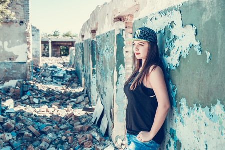 Beautiful, longhaired, dark-haired girl in cap, torn jeans and a black T-shirt on an abandoned house. Attractive woman in jean and cup outfit posing in old ruined factory house. Copy space. Tone photoの写真素材
