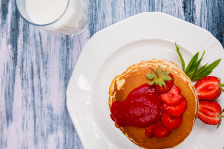 Top view of pancakes with fresh strawberry and jem near glass with milk on white plate on white wooden background.  Stack of pancakes on the table. Copy space.の写真素材