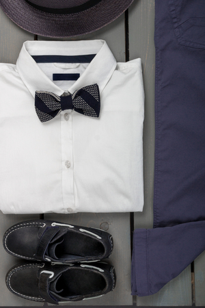 Men's outfit on wooden background. Kids fashion clothes. Grey fedora, navy pant, white shirt, black bow tie and boat shoes for boy. Top view.の写真素材