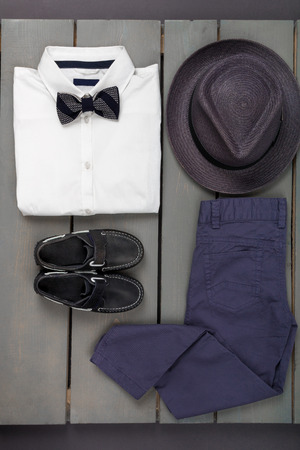 Men's outfit on wooden background. Kids fashion clothes. Grey fedora, navy pant, white shirt, black bow tie and boat shoes for boy. Top view.の写真素材