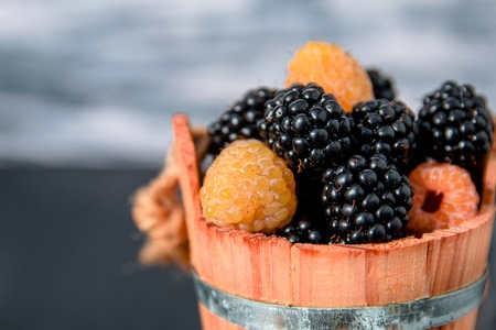 Black and yellow raspberries in a wooden basket on grey wooden background. Close up.の写真素材