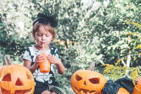 Halloween. Child dressed in black drinking pumpkin cocktail, trick or treat. Little girl near jack-o-lantern decoration in the wood, outdoors.の写真素材