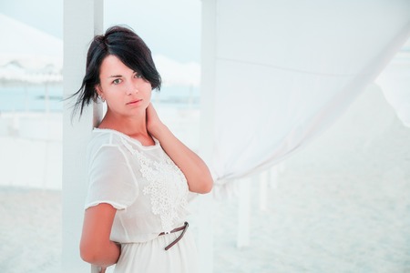 Woman, brunette with short hair in white dress on the beach of ocean, sea near sun shade, tent.の写真素材