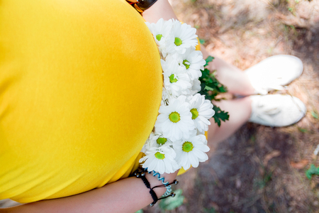 Close up on tummy of pregnant woman, wearing yellow dress, holding in hands bouquet chamomile flowers outdoors, new lifeの写真素材