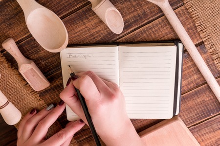 Woman hand write a recipe in cookbook. Book for recipe around utensils on woodenの写真素材