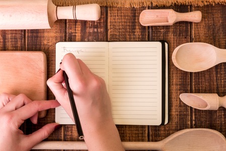 Woman hand write a recipe in cookbook. Book for recipe around utensils on woodenの写真素材