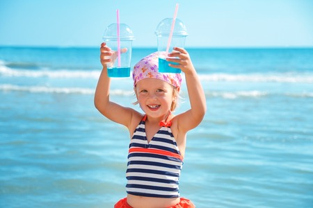 Happy little girl in  swimsuit with two blue cocktail on the beach. Summer. Vacation. Sea. Ocean.の写真素材