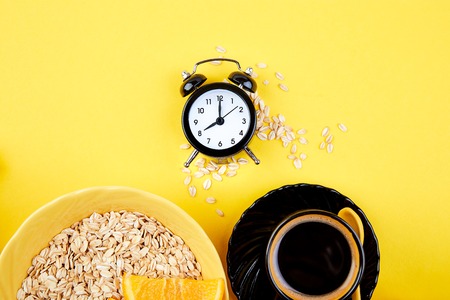 Morning coffee, granola breakfast with fruit near black alarm clock, vase flower on yellow background. Flat lay. Copy space. Top view.の写真素材