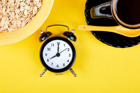 Morning coffee, granola breakfast with fruit near black alarm clock, vase flower on yellow background. Flat lay. Copy space. Top view.の写真素材