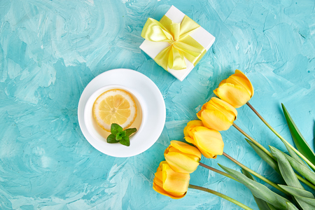 White cup of tea with lemon and gift box near yellow tulip on blue background. Flat lay. Mother or Woman  Day. Greeting Card. Copy space. Spring. Summer. Beautiful.
の写真素材