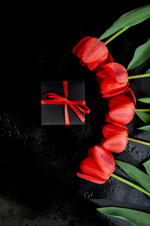 Black gift box with red ribbon near red tulip on black background. Flat lay. Mother or Woman  Day. Greeting Card. Copy space. Spring. の写真素材