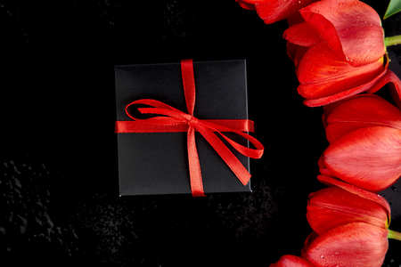 Black gift box with red ribbon near red tulip on black background. Flat lay. Mother or Woman  Day. Greeting Card. Copy space. Spring. の写真素材