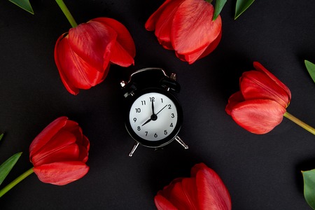 Black alarm clock near bouquet red tulips and cup of lemon tea on black background.Mother or Woman  Day. Greeting Card. Good Morning breakfast. Copy space. Spring. の写真素材