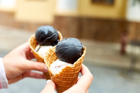 Female hand holding two charcoal and vanila ice cream in waffle cone on city background. Italian traditional ice cream gelato. Copy space.の写真素材
